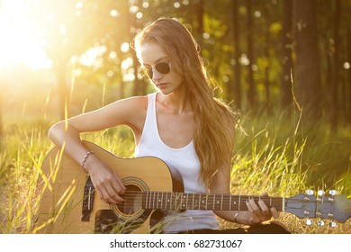 Beautiful female with long luxurious hair wearing white casual shirt and sunglasses playing acoustic guitar while relaxing at greenland enjoying calm atmosphere and beautiful nature. Young musician
