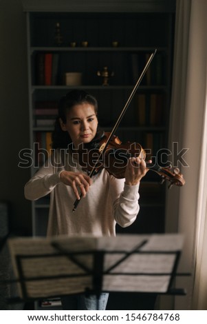 Beautiful female looks at the score while playing the violin. Girl is practicing playing musical instrument at home.