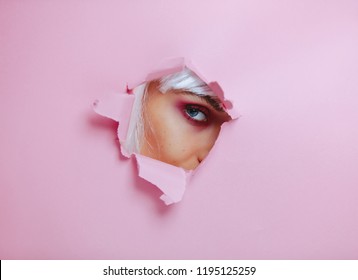 Beautiful Female Model Seeing Through A Hole In Pink Paper.  Woman With Beautiful Eye Makeup Looking Through A Torn Pink Colored Paper.