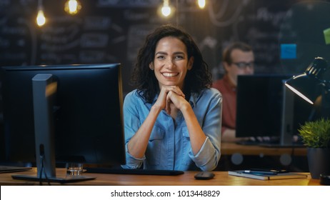 Beautiful Female Office Employee Works At Her Desk On A Personal Computer, She Interrupts Her Work And Smiles Charmingly For The Camera. In The Background  Coworker In The Creative Office.