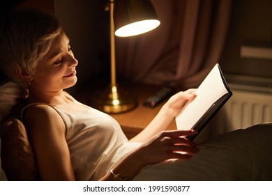 Beautiful female in pajamas lying on bed reading book, alone at night. Caucasian short haired lady in bedroom, charming cute woman in room lighted by lamp. side view