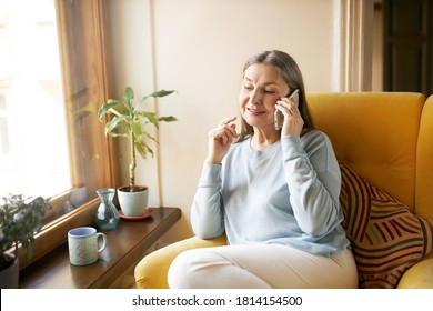 Beautiful female pensioner wearing casual clothes relaxing at home, sitting in armchair by window, drinking morning tea or coffee, smiling, having phone conversation, talking to her old friend - Powered by Shutterstock