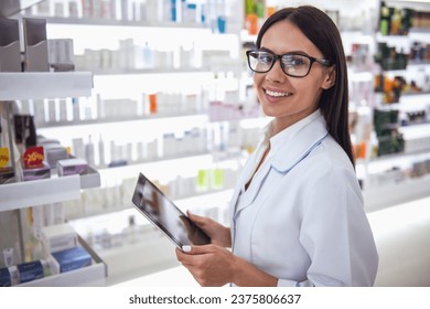 Beautiful female pharmacist is using a digital tablet, looking at camera and smiling while working in pharmacy - Powered by Shutterstock