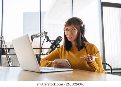 Beautiful female podcaster in headphones and eyeglasses gesturing while recording audio podcast in studio. Young pretty woman in headphones talking into a microphone, holding notepad