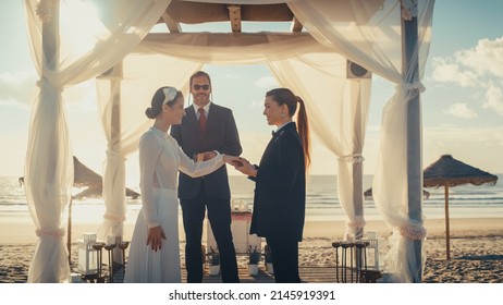 Beautiful Female Queer Couple Exchange Rings And Kiss At Outdoors Wedding Ceremony Near A Sea. Two Lesbian Women In Love Share Their Big Day With Diverse Multiethnic Friends. LGBTQ Relationship Goals.