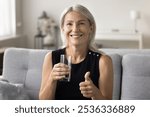 Beautiful female retiree sit on couch look at camera advice recommend you customer drink mineral water for self beauty body care. Senior lady hold glass of aqua propose everyone enjoy pure fresh taste