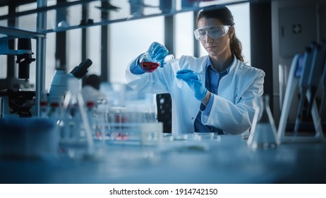 Beautiful Female Scientist Wearing Protective Goggles Mixing Chemicals in a Test Tube in a Lab. Young Professional Microbiologist Working in Modern Laboratory with Technological Equipment. - Powered by Shutterstock