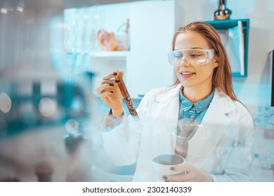 Beautiful Female Scientist Wearing Protective Goggles Mixing Chemicals in a Test Tube in a Lab. Young Professional Microbiologist Working in Modern Laboratory with Technological Equipment.