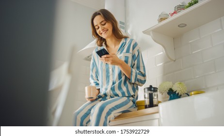 Beautiful Female is Sitting on a Cupboard and Using a Smartphone in a Kitchen While Drinking a Cup of Freshly Brewed Coffee. Young Woman in Pyjamas Smiles When Gets a Text from a Friend. - Powered by Shutterstock