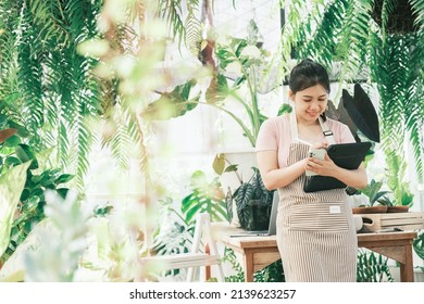 Beautiful Female Smiling Works At Garden Centre Retail Store. Taking Orders Over The Phone. She Working At Table And In Background Is A Shop With Lots Of Plant.