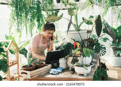 Beautiful Female Smiling Works At Garden Centre Retail Store. Taking Orders Over The Phone. She Working At Table And In Background Is A Shop With Lots Of Plant.