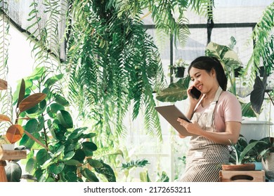 Beautiful Female Smiling Works At Garden Centre Retail Store. Taking Orders Over The Phone. She Working At Table And In Background Is A Shop With Lots Of Plant.