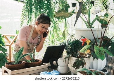 Beautiful Female Smiling Works At Garden Centre Retail Store. Taking Orders Over The Phone. She Working At Table And In Background Is A Shop With Lots Of Plant.