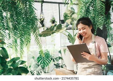 Beautiful Female Smiling Works At Garden Centre Retail Store. Taking Orders Over The Phone. She Working At Table And In Background Is A Shop With Lots Of Plant.