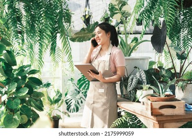 Beautiful Female Smiling Works At Garden Centre Retail Store. Taking Orders Over The Phone. She Working At Table And In Background Is A Shop With Lots Of Plant.