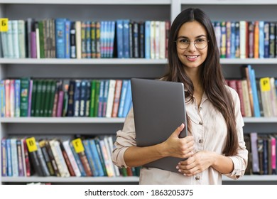 Beautiful Female Student With Laptop Smiling On The Background Of The Library Bookshelves. Learning And Education Online Concept