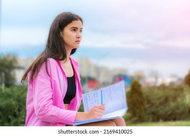 Beautiful Female Student Seated On Bench And Studying At City Street. College Or University Campus Life Style.