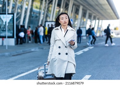 Beautiful Female Tourist Near The Airport, A Chinese Woman Walking With A Big Suitcase, An Asian Woman Holding A Phone, Using The Application For Booking Accommodation And Ordering A Taxi