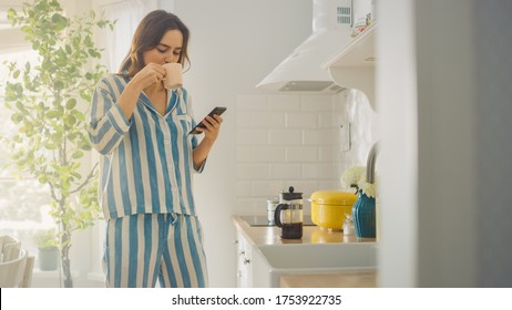 Beautiful Female is Using a Smartphone in a Kitchen While Drinking a Cup of Freshly Brewed Coffee. Girl in Pyjamas with Healthy Lifestyle Relaxes at Home in the Morning. - Powered by Shutterstock