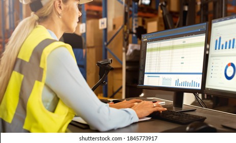 Beautiful Female Worker Wearing Hard Hat Works On Personal Computer Counting Stock In The Retail Warehouse Full Of Shelves With Goods. Commerce, Distribution, Logistics