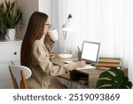 Beautiful female writer with laptop, notebook and cup of tea sitting at table in room