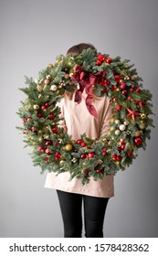 Beautiful Festive Wreath Of Fresh Spruce In Woman Hands. Xmas Circlet With Red And Gold Ornaments And Balls. Christmas Mood. Gray Wall On Background.