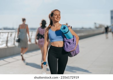 Beautiful fit caucasian girl in sportswear walks at embankment carrying yoga mat bottle of water looks at camera tooth smiling. Cheerful American young woman goes to exercise at beach. - Powered by Shutterstock