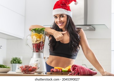 Beautiful Fit Female In Christmas Hat Leans Against The Blender Full Of Healthy Fruit And Veg In A Blender.