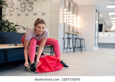 Beautiful fit sportswoman looking at camera smiling sitting on bench tying her shoelaces in modern bright locker room getting ready to exercise workout in gym and keep her body healthy and in shape. - Powered by Shutterstock