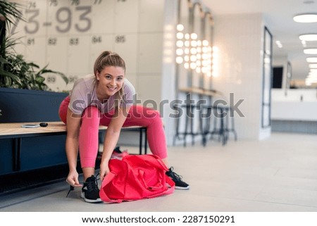 Beautiful fit sportswoman looking at camera smiling sitting on bench tying her shoelaces in modern bright locker room getting ready to exercise workout in gym and keep her body healthy and in shape.