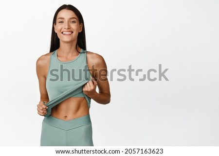 Beautiful and fit sportswoman showing her abs, smiling and enjoying results of workout in gym, standing over white background
