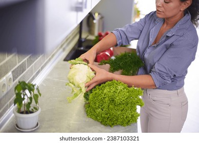 Beautiful fit woman in casual wear, standing by kitchen counter and taking fresh organic vegetables and greens out of a cardboard box, delivered at home. Housewife sorting groceries for cooking meal - Powered by Shutterstock