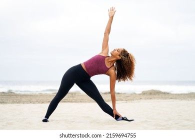 Beautiful fit young African American woman in yoga triangle pose or Trikonasana at the beach wearing workout clothing and stretching                                         