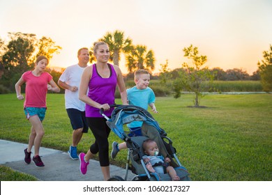 Beautiful, Fit Young Family  Jogging Together Outdoors Along A Paved Sidewalk In A Park Pushing A Stroller At Sunset