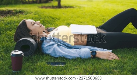 Beautiful fitness athlete woman relaxing on green grass while reading a book after exercising, taking rest after workout outdoor. Healthy concept. ealthy