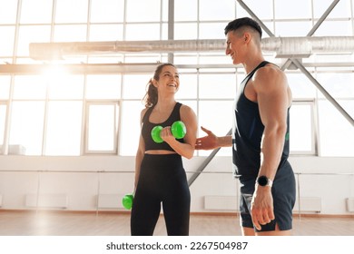 beautiful fitness girl in sportswear trains with a trainer in bright gym, man trainer helps woman lift dumbbells at fitness training - Powered by Shutterstock