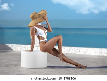 Beautiful fitness woman with perfect butt and legs in white swimwear and large hat posing on the white-colored Santorini resort shore.