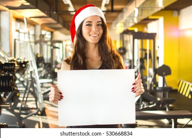 Beautiful fitness woman in Santa hat holding empty board. New Year. Christmas, holidays, fitness, and gym concept.