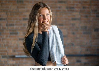 Beautiful Fitness Woman With A Towel Eating A Protein Bar After Working Out At The Gym. Sporty Attractive Woman After Training. Sport And Recreation Concept.