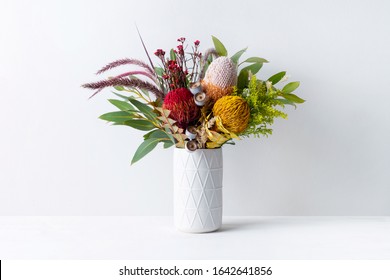 Beautiful Floral Arrangement Of Mostly Australian Native Banksias, Eucalyptus Leaves And Gum Nuts, Fountain Grass And Dried Kangaroo Grass, In A White Vase On A White Table With A White Background.