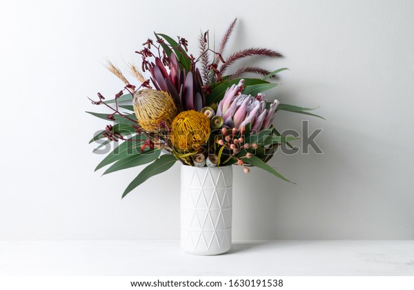 Beautiful floral arrangement of mostly\
Australian native flowers, including protea, banksia, kangaroo paw,\
eucalyptus leaves and gum nuts, in a white vase on a white table\
with a white\
background.
