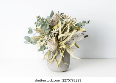 Beautiful flower arrangement of Australian native dried banksia, eucalyptus leaves and delicate white flowers, in a grey vase on a table with a white background. - Shutterstock ID 2275792223
