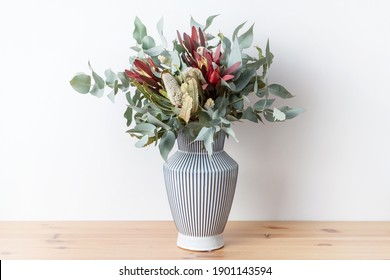 Beautiful flower arrangement of mostly Australian native flowers, including Banksia, Silvan Reds and Eucalyptus leaves, in a white and blue strip vase, with a white background on a wooden table. - Shutterstock ID 1901143594