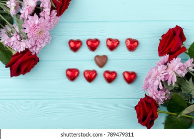 Beautiful Flowers And Heart Shaped Chocolate. Valentines Day Flowers And Chocolate Hearts On Wooden Background, Top View. Happy Valentines Day.