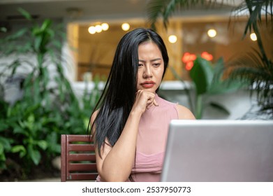 A beautiful, focused Asian woman is working remotely, working on her laptop at an outdoor cafe, looking at her laptop screen with a thoughtful face. people, lifestyles, wireless technology - Powered by Shutterstock