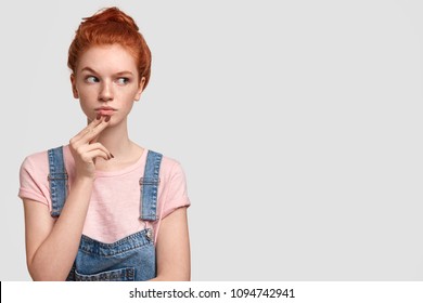 Beautiful freckled female with ginger hair bun, raises eyebrow in bewilderment, looks suspiciously aside, notices something strange, holds chin, wears denim overalls, poses against white wall