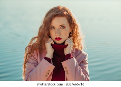 Beautiful freckled readhead woman with natural long curly hair, bold marsala color lips makeup, wearing cashmere turtleneck sweater, pink blazer. Copy, empty space for text