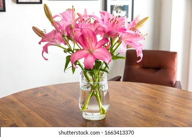 Beautiful fresh pink lilies with a glass jug as a vase on a nice wooden acacia table with a cognac leather lounge chair against a white wall with photos. 