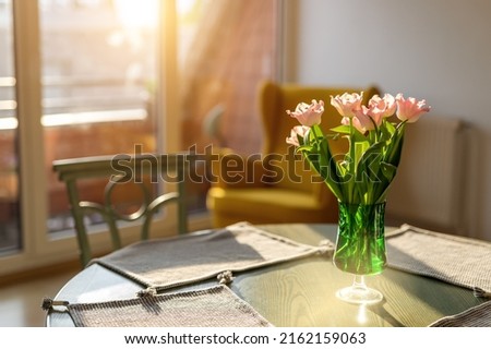 Beautiful fresh pink tulips bouquet in green glass vase on table in warm sunset sun lights against balcony window in cozy home interior. Blooming flowers decoration in living room provence style