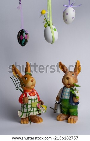 Beautiful front view at two easter bunnies, boy and girl bunny, with gardening stuff in their hands, looking up on three painted easter eggs above their heads, with white isolated background.